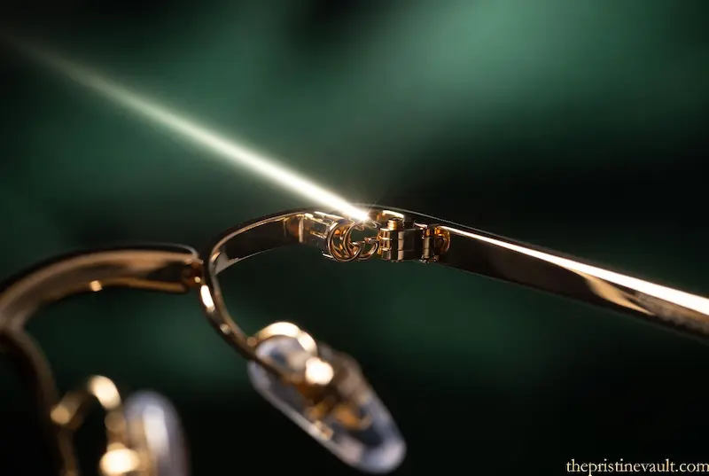 Extreme close-up of a genuine gold Gucci eyeglass hinge with a brilliant light reflection, showcasing the precise interlocking GG logo and premium construction.