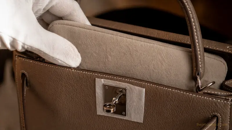 A professional specialist wearing white gloves inserting a velvet bag shaper into an Etoupe Hermès bag, with acid-free tissue paper protecting the leather behind the hardware latch.