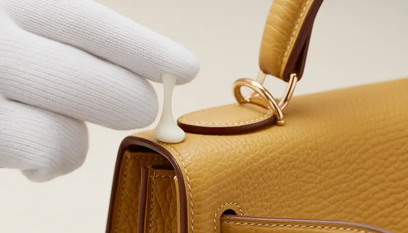 A specialist in white gloves applying a pea-sized amount of Saphir Renovateur to a grained Hermès Togo bag.