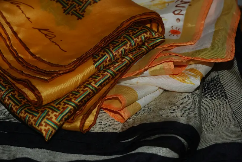 Multiple Hermes silk scarves in gold, orange, and ivory prints folded and stacked on top of each other, showing the rich silk texture and intricate woven border details