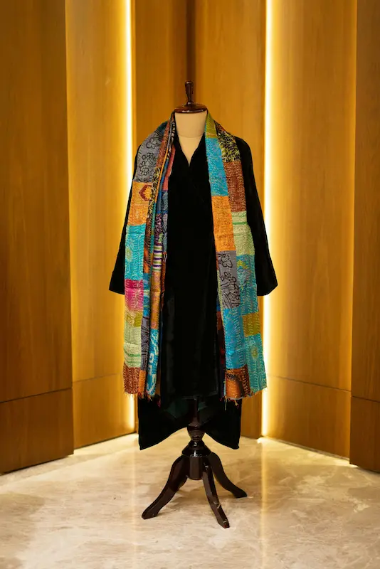 A colorful Hermes silk scarf with vibrant multicolor patchwork print draped over a black dress mannequin, displayed against warm amber-lit wood panel walls