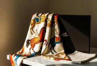 A Hermes silk scarf with equestrian and horseman print draped over a marble surface beside the original black Hermes box and ivory dust bag, lit by soft natural side lighting