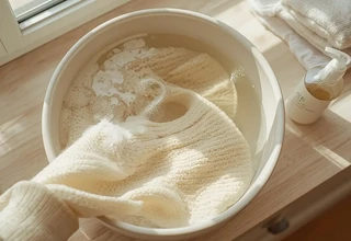 Hand washing a delicate cream cashmere sweater in a white basin filled with soapy water next to a gentle liquid detergent