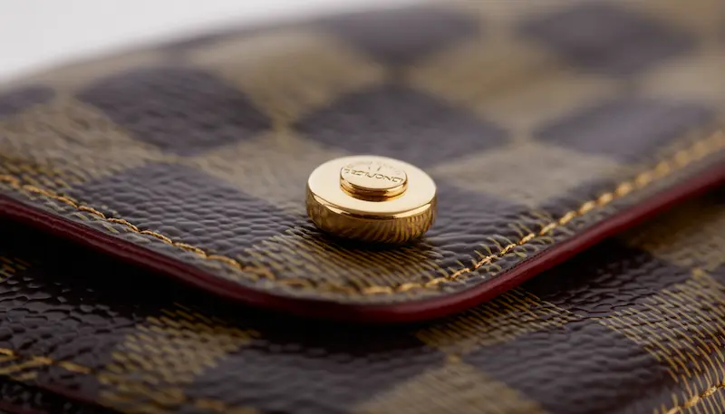 Macro shot of the gold-plated snap button on a Louis Vuitton Victorine Wallet in Damier Ebene canvas, showing detailed stitching and the circular impression point on the canvas.
