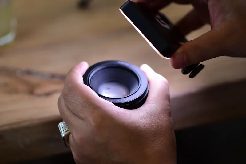 Hands inspecting a Canon EF camera lens elements with a portable LED light to check for fungus or mold growth